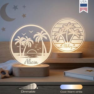 May include: Two illuminated, round night lights with a beach scene design, featuring palm trees, a sunset, and the names "Alicia" and "Abby." The lights sit on wooden bases. The background includes a crescent moon and stars. "Made in USA" is printed on the top right.