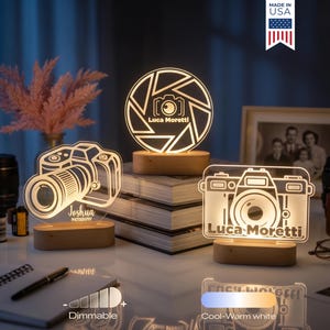 May include: Three illuminated camera-themed night lights with wooden bases. One features a camera lens design, another a camera outline with "Luca Moretti", and the third a camera outline with "Joshua". The lights are dimmable and offer cool-warm white light.