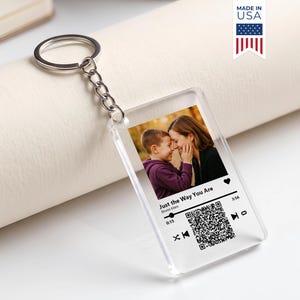 May include: A rectangular clear acrylic keychain featuring a printed photograph of a child and an adult. The design includes a music player interface with the text Just the Way You Are by Bruno Mars, a heart icon, a QR code, and playback controls.