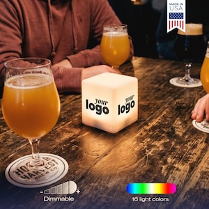May include: A lit cube with the text "your logo" on each side sits on a wooden table with beer glasses. The image shows a dimmable light with 16 light colors. A coaster with "HO HARVEST BREW CO" is visible.