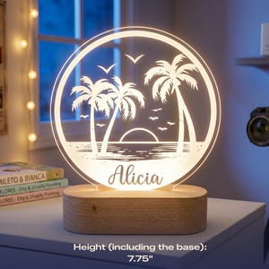 May include: A personalized LED night light featuring a beach scene with palm trees, seagulls, and a sunset. The name "Alicia" is displayed below the image. The light sits on a wooden base and measures 7.75 inches in height.