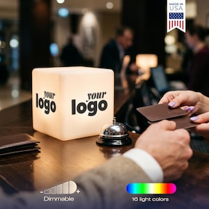 May include: A glowing white cube with the words "your logo" printed on two sides. The cube sits on a dark wooden surface with a wallet and a bell. The image also shows a color spectrum and the text "16 light colors".