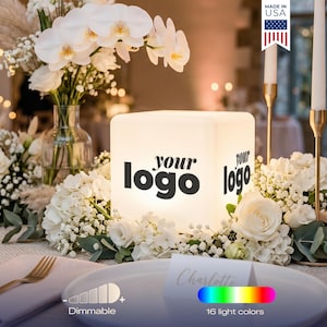 May include: A white illuminated cube with "your logo" printed on it, surrounded by white flowers and candles. The cube has dimmable settings and 16 light colors. "Made in USA" is printed on a flag.