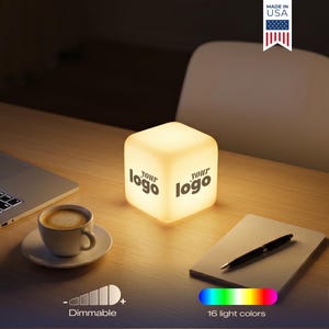 May include: A glowing cube-shaped lamp with the text "your logo" on two sides. The lamp is on a wooden desk with a laptop, coffee cup, notepad, and pens. The lamp is dimmable and offers 16 light colors. Made in the USA.