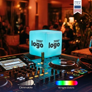 May include: A DJ setup with a glowing, dimmable, light-up cube displaying "your logo" in black. The cube emits a soft blue light and sits atop a DJ mixer with colorful lights. The image also shows a wine glass and headphones.