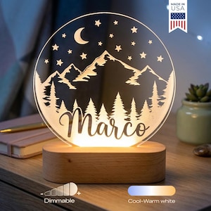 May include: A personalized night light with a mountain and forest design, a crescent moon, and stars. The name "Marco" is displayed in a script font. The lamp sits on a wooden base and is dimmable.