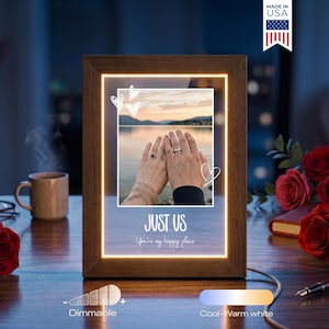 May include: A rectangular wooden frame with an integrated LED light border surrounding a central photograph of two hands wearing rings against a lake sunset. The frame features the text JUST US and You're my happy place, with hand-drawn heart icons.