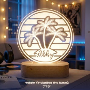 May include: A personalized night light with a circular design featuring palm trees and the name "Abby." The light is set on a wooden base and measures 7.75 inches in height. The light emits a warm glow, perfect for a bedroom or living space.