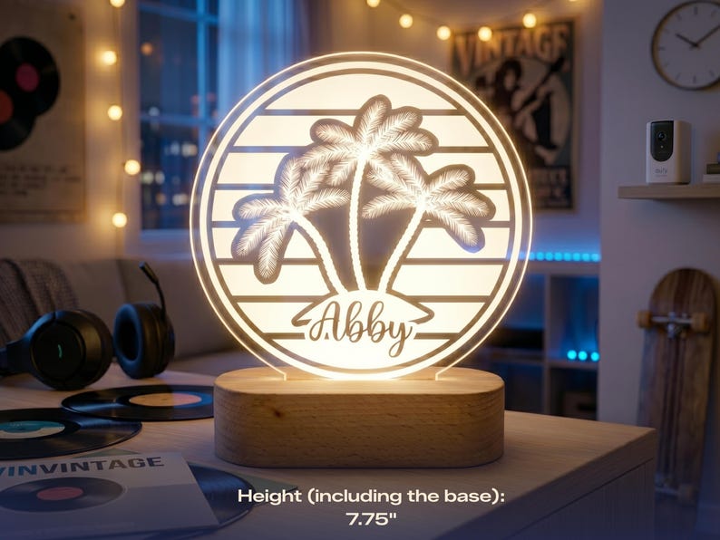 May include: A personalized night light with a circular design featuring palm trees and the name "Abby." The light is set on a wooden base and measures 7.75 inches in height. The light emits a warm glow, perfect for a bedroom or living space.