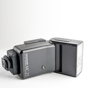 May include: A black Cullmann MC25 SCA 300 dedicated camera flash unit. The flash has a rectangular body with a flash head on the right side and a mounting foot on the left. The text "CULLMANN MC25 SCA 300 dedicated" is printed on the side.