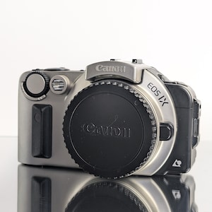May include: A silver and black Canon EOS IX camera with a lens cap. The camera has the Canon logo and EOS IX model name printed on it. The camera is on a reflective surface.