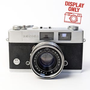 May include: A vintage Konica Hexanon camera with a silver top and black body. The lens is labelled "Konica Hexanon 45mm 1:1.8". The camera has a "New Loading System" sticker and a "Display Only" stamp.