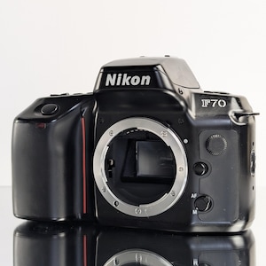 May include: Black Nikon F70 camera with a silver lens mount. The camera has the Nikon logo in white on the top and the model number F70. A red stripe accents the side of the camera.