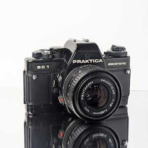 May include: A black Praktica BC 1 electronic 35mm film camera featuring a Pentacon Prakticar 1:1.8 f=50mm MC lens. The camera body includes various dials, a shutter release button, and a hot shoe mount on top, with the brand name clearly visible.