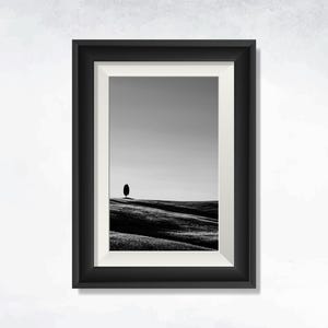 Puede incluir: Impresión enmarcada en blanco y negro de un paisaje. La obra presenta un árbol solitario en una colina ondulada bajo un cielo despejado. El marco negro con un paspartú blanco crea una estética clásica y minimalista.