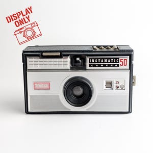 May include: A vintage Kodak Instamatic 50 camera with a black and silver body. The camera features the text "Instamatic 50 Camera" and the Kodak logo. A red stamp in the top left corner reads "DISPLAY ONLY".