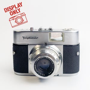 May include: A vintage Voigtländer camera with a silver body and black leather accents. The lens is labelled "COLOR-SKOPAR 1:2.8/50". The camera has a flash and various dials. The words "DISPLAY ONLY" are stamped in red.