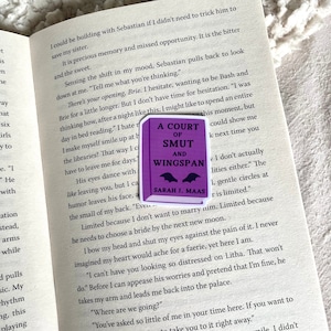 Op de afbeelding: Een close-up van een boek met een paarse sticker op de kaft. De sticker toont een zwarte vleermuis met witte vleugels en de tekst "A Court of Smut and Wingspan" in wit. De tekst "Sarah J. Maas" staat in wit onder de titel. Het boek is open op een pagina met tekst over een personage genaamd Brie.