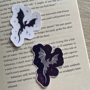Op de afbeelding: Twee zwart-witte draken stickers met een sterrenhemel achtergrond. De draken vliegen met hun vleugels uitgespreid. De stickers zijn op een boekpagina met tekst over een koningin.