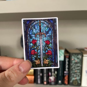 Op de afbeelding: Een sticker van glas-in-lood met een zwaard met een gouden handvat en een blauw, rood en geel glas-in-lood ontwerp. Het zwaard is omringd door rode rozen en gele bloemen.