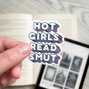 Op de afbeelding: Een sticker met de tekst "HOT GIRLS READ SMUT" in een regenboogkleurenschema. De sticker zit op een boek met een zwart-witte e-reader op de achtergrond.