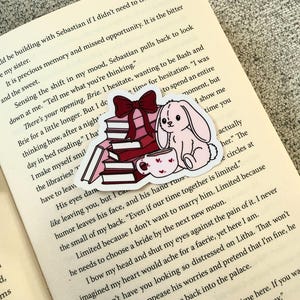 Pode incluir: Um autocolante com uma pilha de livros com um laço vermelho, um coelho rosa e uma chávena de chá com corações. O autocolante está colocado num livro aberto. O tema é leitura e literatura, com um design fofo.