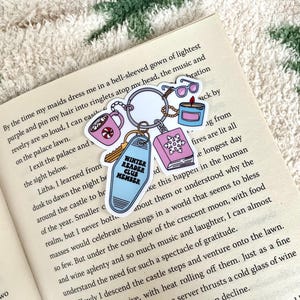 Op de afbeelding: Een sleutelhanger met diverse bedels, waaronder een mok, zonnebril, kaars, boek en een sleutelhanger met de tekst "Winter Reader Club Member". De items zijn in pastelkleuren en liggen op een open boek.