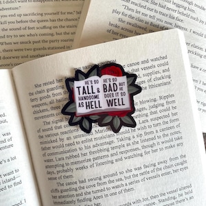 Könnte beinhalten: Ein schwarz-roter, gestanzter Aufkleber mit dem Text "HE'S SO TALL & BAD BUT HE HANDSOME DOES IT SO AS HELL WELL" auf weißem Hintergrund, umgeben von einem floralen Design. Der Aufkleber befindet sich auf den offenen Seiten eines Buches.