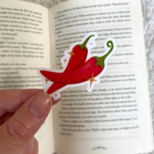 Op de afbeelding: Sticker met een rode chilipeper, een witte omtrek en gouden glitteraccenten. De sticker zit op een boek met tekst.