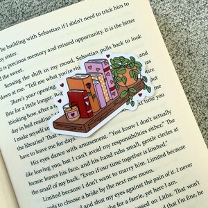 Op de afbeelding: Een sticker met een boekenplank met boeken, een plant in een pot en een kaars. De boeken zijn in verschillende kleuren, en kleine hartjes zijn verspreid over het ontwerp. De sticker is geplaatst op een open boek.