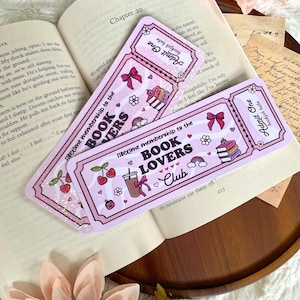 Op de afbeelding: Twee roze "Book Lovers Club" bladwijzers met ticketontwerpen, met kersen, strikken en een drankillustratie. De bladwijzers liggen op een open boek met hoofdstuk 20.
