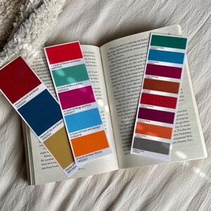 Op de afbeelding: Een set van tien kleurrijke bladwijzers met verschillende tinten rood, oranje, geel, groen, blauw en bruin. Elke bladwijzer heeft een titel en een datum erop gedrukt. De titels omvatten "House of Earth and Blood", "A Court of Thorns and Roses", "A Court of Mist and Fury", "A Court of Wings and Ruin", "A Court of Frost and Starlight", "A Court of Silver Flames", "The Assassin's Blade", "Crown of Midnight", "Empire of Storms" en "Tower of Dawn".