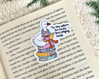 Op de afbeelding: Een witte sticker met een cartoon gans met een sjaal en een mok, zittend op een stapel boeken. De sticker heeft de tekst "'Tis the season for books and staying inside."