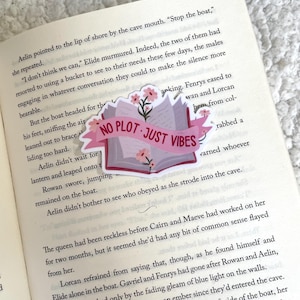 Op de afbeelding: Een boek met een roze en witte omslag met de tekst "No Plot - Just Vibes". Het boek is open en heeft een roze bloem bovenop de pagina. Het boek ligt op een pagina van een boek met tekst over een boot en een grot.