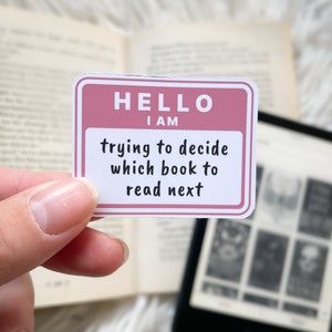 May include: A pink and white sticker with the text "HELLO I AM trying to decide which book to read next".