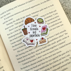 Op de afbeelding: Een gestanste sticker met de tekst "I love lil snacks and paperbacks" en illustraties van een croissant, matcha, een boek, cake en bubble tea. De sticker ligt op een open boek.