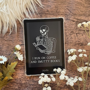 Op de afbeelding: Een zwart-witte illustratie van een skelet dat een boek en een kop koffie vasthoudt. De tekst luidt "Ik draai op koffie en smerige boeken." De afbeelding wordt weergegeven op een Kindle e-reader.