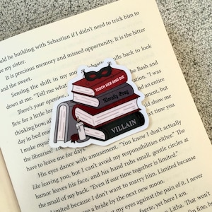 Op de afbeelding: Een sticker met een stapel boeken met de titels "Touch Her and Die", "Morally Gray" en "Villain". Een zwart masker en een kaars zijn ook afgebeeld. De sticker is op een open boek geplaatst.