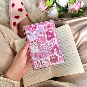 May include: A pink tablet case decorated with stickers, including a swan, lips, and the words "Bye" and "XOXO." The case is held over an open book. A pink slipper with red hearts is visible.