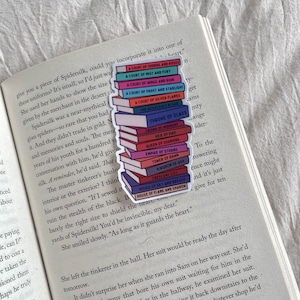 Könnte beinhalten: Ein farbenfroher Aufkleber mit einem Stapel von Büchern mit Titeln wie "A Court of Thorns and Roses" und "Throne of Glass". Der Aufkleber befindet sich auf einem offenen Buch mit gedrucktem Text und erzeugt ein literarisches Thema.
