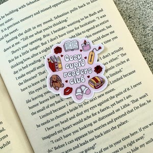Peut inclure: Un autocollant rose avec le texte "Cozy Cupid Readers Club" et des illustrations de livres, une bougie, une étiquette, des roses, un lapin, des pantoufles et une couverture. L'autocollant est sur un livre ouvert.