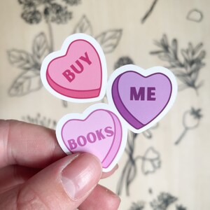 Op de afbeelding: Drie roze en paarse hartvormige stickers met de woorden "KOOP", "MIJ" en "BOEKEN" erop.