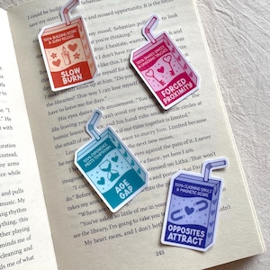Puede incluir: Una colección de pegatinas coloridas con forma de caja de zumo con pajitas, colocadas sobre un libro abierto. Cada pegatina presenta texto y gráficos, incluyendo "Slow Burn", "Forced Proximity", "Age Gap" y "Opposites Attract".