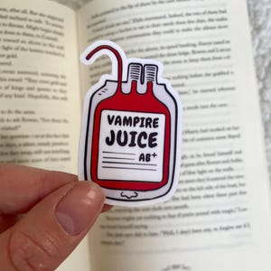 Op de afbeelding: Een witte sticker in de vorm van een bloedzak met de woorden "VAMPIRE JUICE AB+" in het zwart. De zak is rood met een witte omtrek en heeft een gebogen buis aan de bovenkant. De sticker wordt voor een open boek gehouden.