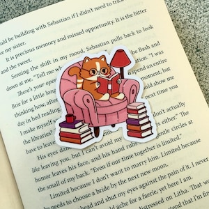 Può includere: Un adesivo raffigurante un gatto dei cartoni animati con gli occhiali che legge un libro in una poltrona rosa, con una lampada rossa e pile di libri. L'adesivo è posto su un libro aperto.