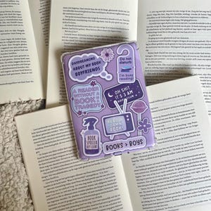 以下が含まれることがあります： 紫と白のブックカバーに、さまざまなステッカーが貼られています。ステッカーには、「Daydreaming About My Book Boyfriends」、「A Reader Without a Book is a Tragedy」、「Read a Damn Book」、「Oh Shit It's 3 AM Book Club」、「Book Spoiler Repellent」、「Do Not Disturb: I'm Busy Reading!」などのテキストが書かれています。
