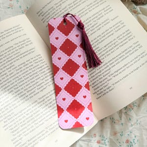 Op de afbeelding: Een decoratieve boekenlegger met een rode achtergrond met een herhalend patroon van roze vierkanten met geschulpte randen en kleine roze hartjes. Een bordeauxkleurige kwast hangt eraan. De boekenlegger ligt in een open boek.