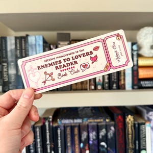 Op de afbeelding: Een roze en witte bladwijzer met de tekst "lifetime membership to the ENEMIES TO LOVERS READER Book Club." De bladwijzer heeft illustraties van een hart, lippen en een gebroken hart. De woorden "Admit One" zijn ook zichtbaar.