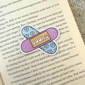 Peut inclure: Un autocollant en forme de pansement avec le mot "BOOKS" dans un rectangle jaune. Le pansement a une section bleu clair avec des fleurs blanches et une section violette avec de petits points.