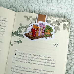 Può includere: Un segnalibro colorato con una libreria in miniatura con libri, una candela e una pianta in vaso. Il segnalibro è posto in un libro aperto, con il numero di pagina 24 visibile. Il segnalibro ha una linguetta marrone.